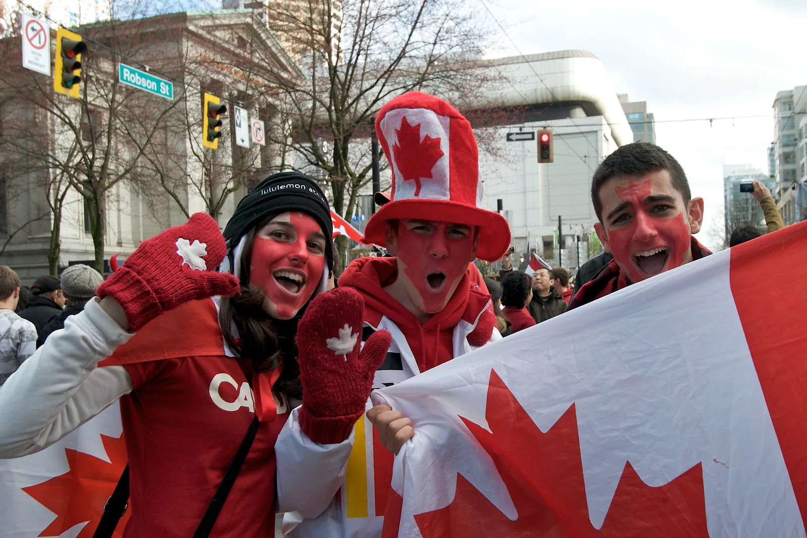 Three smiling sports fans with red and white face paint and a flag, photographed in daylight on a city street