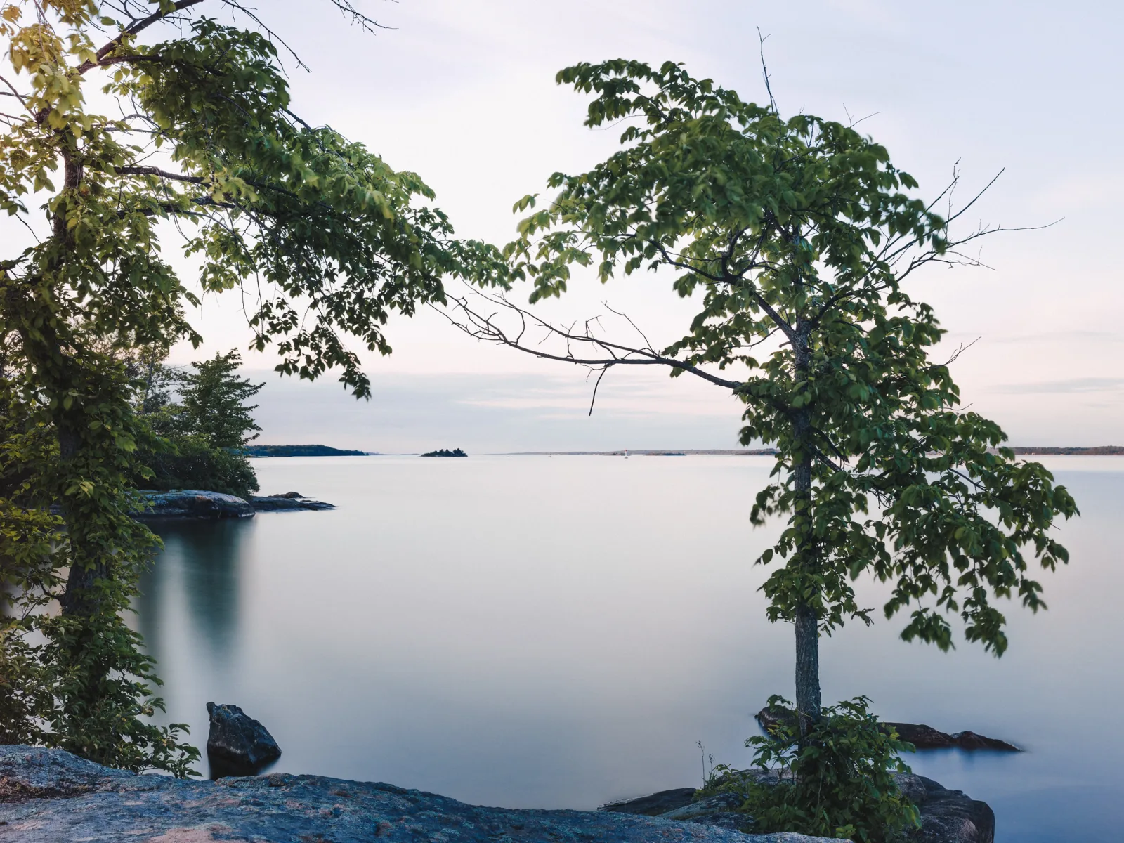 Trees on a rocky shore frame a wide stretch of water made smooth and glassy by a long exposure, under a pale sky.