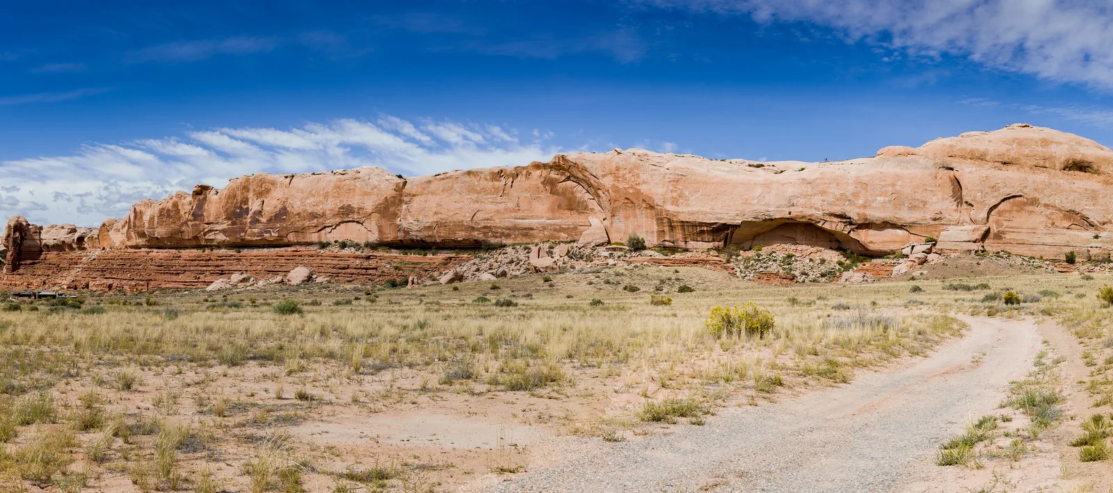 A dirt track leading toward a long sandstone ridge with small arches under a deep blue sky