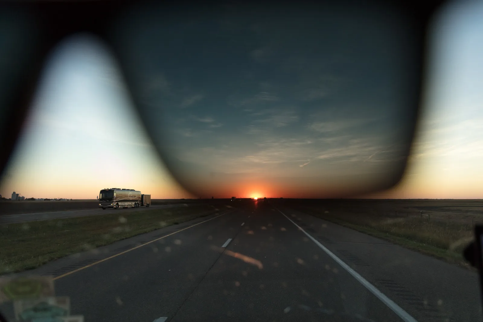 A road at sunrise seen through the dark lens of a pair of sunglasses, with the edge of the frame blurred in the foreground