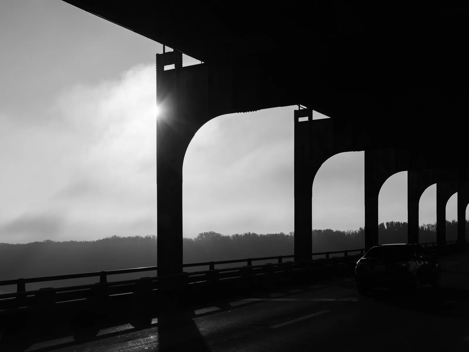 Black and white view from a highway bridge deck, with dark steel arches framing a misty river valley and a single car driving across