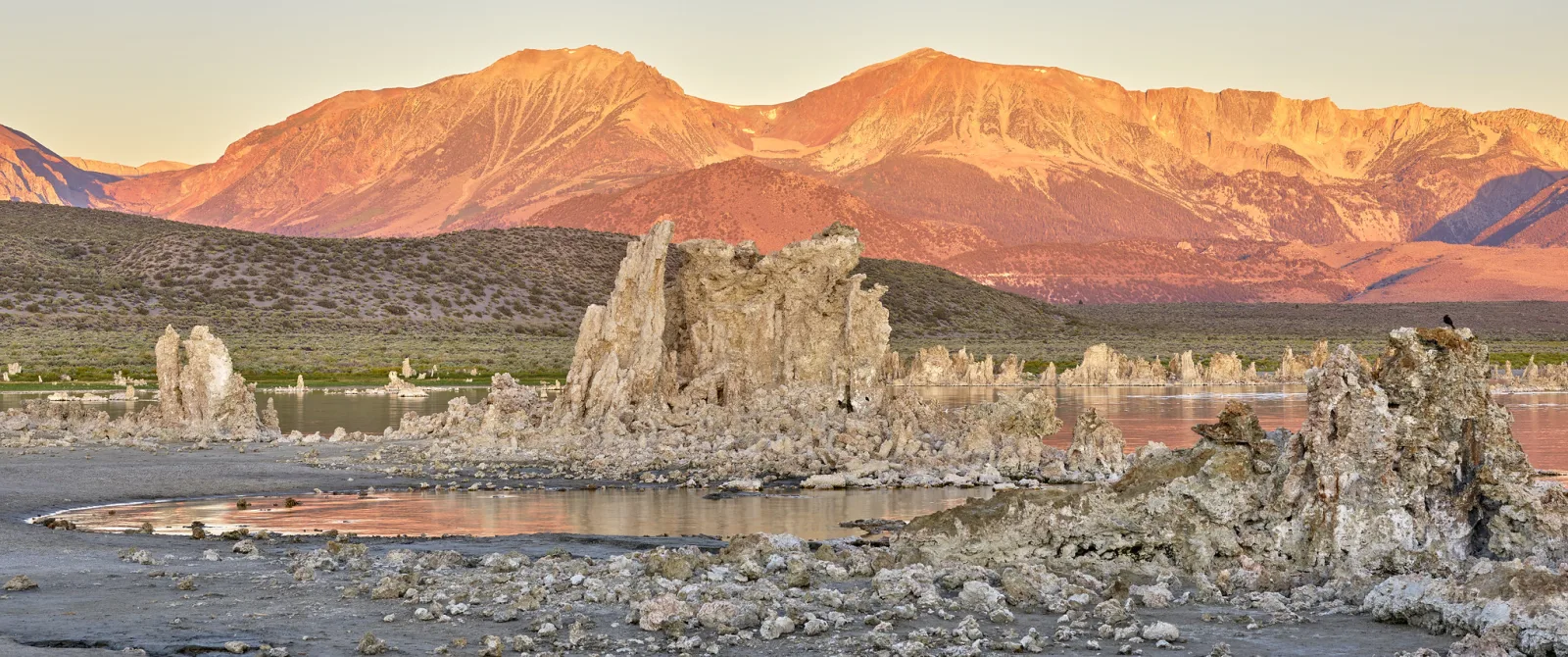 Rock tufa towers at the edge of a lake with orange sunrise light on the mountains behind.