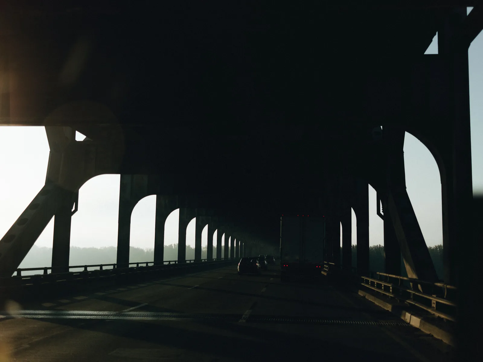 Looking along the dark underside of a multi-level elevated bridge, its heavy pillars receding toward a band of daylight.