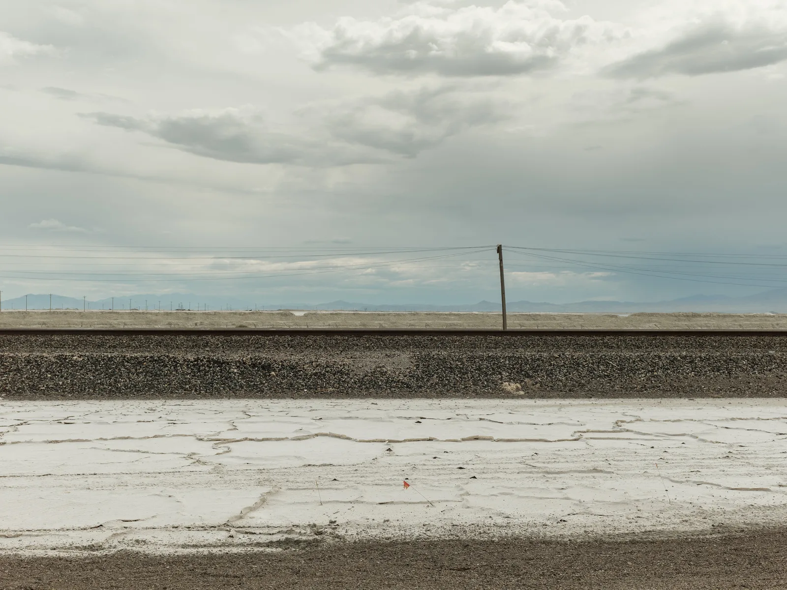 Cracked white salt crust and dark gravel beside a railway line under a pale cloudy sky, with distant mountains