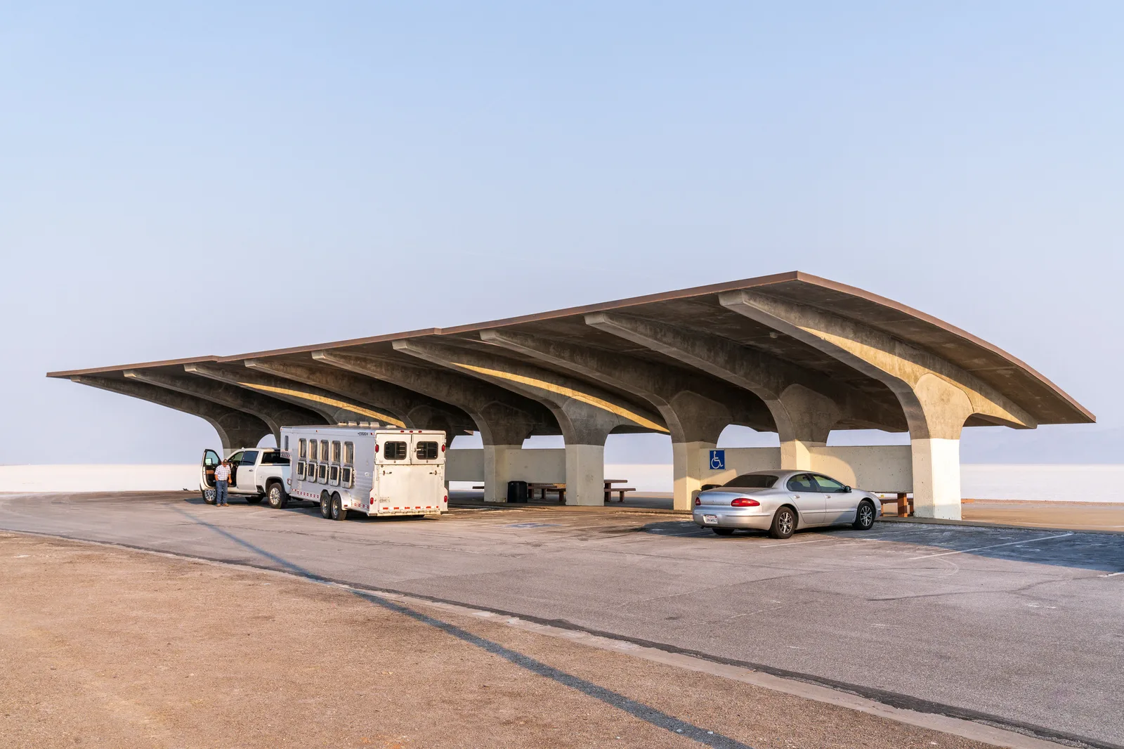 A curved concrete shelter at a rest area beside a pale, bright salt flat under a soft blue sky