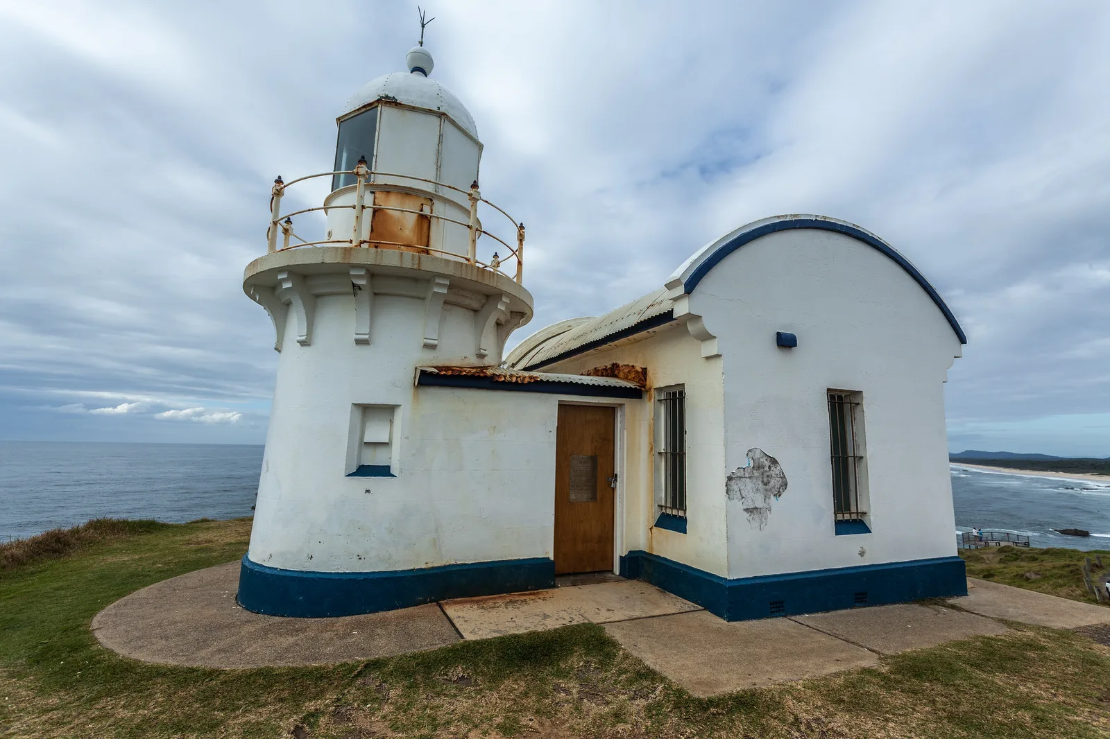 A small white lighthouse with blue trim and an attached domed building on a grassy headland above the sea under a cloudy sky.